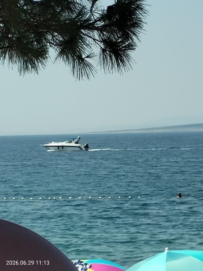 An elevated, slightly angled, medium shot captures a tranquil scene by the sea in Krk, Croatia. The foreground is partially obscured by the dark, shadowy branches of a pine tree, its needles sharp and defined against the pale, hazy sky. Beyond the tree, the deep blue sea stretches out, disturbed by gentle ripples. A white motorboat with a blue canopy navigates the water, leaving a white wake in its path. In the mid-distance, a person is swimming in the sea, their head barely visible above the surface. A line of white buoys floats on the water, marking a boundary. In the far distance, a thin strip of land, possibly a coastline, is visible as a faint, hazy outline against the horizon. The lower portion of the image reveals a portion of a beach umbrella and a dark-colored beach chair, suggesting a sunny day at the shore. The date stamp "2026.06.29 11:13" is visible in the bottom left corner of the image.
