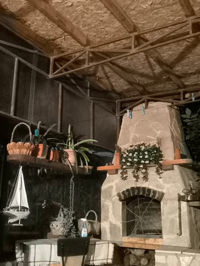 The image depicts an outdoor covered area, possibly a patio or veranda, at night. The structure has a wooden beam ceiling with plywood sheeting and exposed metal framing. To the right, a large stone fireplace or oven is prominent. It has a decorative shelf with a floral arrangement of small white flowers and greenery. Below the shelf, there is a rectangular opening with a large, circular metal fan grill. The area appears to be a domestic or restaurant setting in Kichevo, Bulgaria.

On a wooden shelf mounted on the wall to the left, there are three potted plants and a woven basket. Below this shelf, a model sailboat is displayed, resting on a table covered with a checkered cloth. A metal chain hangs from above, attached to a decorative object that appears to be made of metal or wire. Next to the chain, there is a bottle of cleaning spray and another woven basket. A chair is partially visible in the foreground. The overall impression is of a rustic, somewhat cluttered outdoor space, possibly used for dining or entertaining.