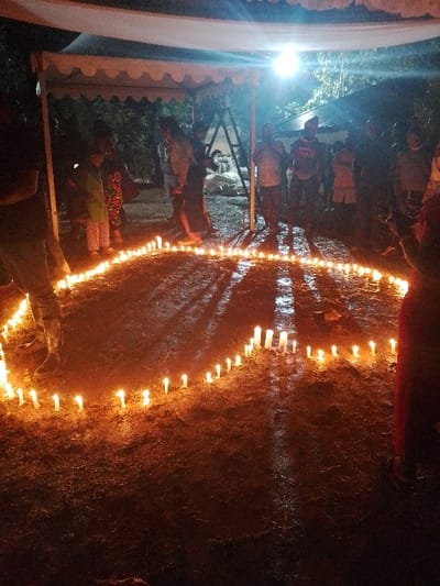 The scene depicts a nighttime outdoor gathering in Marenyo-Shianda ward, Kenya. Approximately fifteen individuals, including adults and children, are standing around a large, elongated, and irregularly shaped perimeter marked by numerous lit white pillar candles placed directly on damp, dark earth. A white fabric canopy with a scalloped edge is partially visible in the upper-left, supported by metal poles; a bright, overhead electric light source is positioned beneath this canopy, casting strong illumination and creating distinct reflections and shadows on the wet ground.
In the immediate foreground, a person on the right, wearing a red garment, is partially visible with both arms raised. On the left, another individual is partially visible from the waist down, wearing dark trousers and boots. Other individuals are distributed in the mid-ground and background, observing the central candle arrangement. Some individuals are standing directly under the canopy. The background is dimly lit, revealing dark foliage and an indistinct structure, possibly another tent frame. The wet ground conditions and collective focus on the illuminated candle arrangement suggest a structured outdoor event or vigil.
