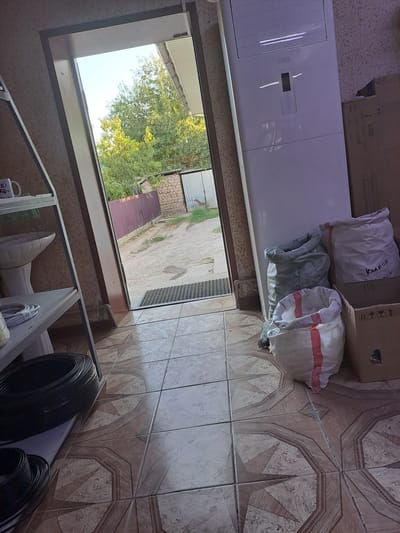 The image depicts an interior scene looking out through an open doorway into an outdoor area. Inside, on the left, is a white shelving unit with a white sink and some white coiled cables. In the foreground, there are large black coiled cables. On the right, there is a tall white floor-standing air conditioner unit. Next to the air conditioner, there are three large sacks, two of which are white with red stripes and one is a gray mesh bag. A brown cardboard box sits to the right of the sacks. The floor is tiled with a patterned design in shades of brown and beige. The outdoor area visible through the doorway appears to be a yard with a dirt path, a purple corrugated metal fence, and trees with green foliage. The sky is visible and appears to be clear and blue. The scene suggests a storage or utility area. The location is identified as Асықата кенттік әкімдігі, Kazakhstan.
