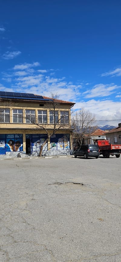 The image shows a street scene in Slatino, Bulgaria. A two-story yellow building occupies the left side of the frame, adorned with blue advertisements for ice cream. Solar panels are visible on the roof. A bare tree stands in front of the building. A black car and a red truck are parked on the right side of the frame, along the edge of a gray asphalt road. A clear blue sky with scattered white clouds fills the upper left portion of the image. The image appears to be taken on a sunny day.