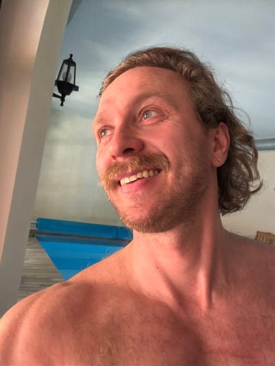 A close-up, angled selfie of a shirtless man with long, wavy brown hair, a brown beard and mustache, and light eyes. He has a small earring in his left ear. The background is a pale blue wall with a black lantern fixture and a blue cabinet. The location is Тэльмінскі сельскі Савет, Belarus.