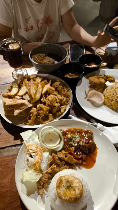 The image depicts a dining scene at a restaurant in Berastagi, Indonesia. A person, wearing a white t-shirt with a graphic print, is visible in the upper portion of the frame. They are holding a smartphone in their right hand, engaged with it. The table is laden with various food items and drinks.
In the foreground, a white plate holds a meal consisting of white rice topped with a fried egg or similar item, a portion of fried chicken pieces, a side salad with shredded carrots and lettuce, and a piece of meat covered in a dark, glossy sauce, garnished with chopped green onions and red pepper slices, accompanied by a lemon wedge.
To the left of this plate is another white plate filled with a generous serving of fried calamari rings and potato wedges. Adjacent to this plate, a tall glass contains a dark beverage, possibly iced coffee or tea, with a slice of lemon.
Further back on the table, a dark bowl contains what appears to be a saucy dish with some fried elements. Two smaller black bowls hold dipping sauces, one appearing to be a yellowish-brown sauce and the other a reddish sauce, possibly chili sauce or ketchup. In the background, a smartphone is placed on the table.
The overall environment suggests an indoor dining setting with wooden table surfaces, illuminated by ambient lighting. The focus is on the food and the act of dining, with a casual and relaxed atmosphere.