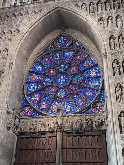The image shows a large, intricately designed rose window above a pair of massive wooden doors, part of a grand stone building, likely a cathedral or church. The rose window is filled with vibrant stained glass, predominantly in shades of blue, red, and yellow, forming geometric patterns and figural scenes. Surrounding the rose window and on the facade of the building are numerous stone sculptures of figures, arranged in niches and rows. These figures appear to be religious in nature, possibly saints or biblical characters, depicted in various poses and with distinct clothing. The wooden doors are imposing and appear to be ancient, with a rustic, planked design. The overall impression is one of significant architectural and artistic detail, indicative of a historical and possibly religious site. The location is identified as Urvillers, France, though without further context or specific landmark identification within Urvillers, the exact building cannot be pinpointed.