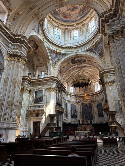 The image depicts the interior of a grand church, likely a cathedral, featuring opulent Baroque or Rococo architecture. The dominant colors are white and gold, with intricate decorative patterns adorning the walls, columns, and ceilings. A large dome is visible at the top, adorned with frescoes and illuminated by natural light from windows. Several large paintings hang on the walls, depicting religious scenes.
The foreground shows rows of dark wooden pews, some occupied by individuals. The floor is a checkered pattern of black and white tiles. In the mid-ground, there are pulpits and architectural elements typical of a church, including an altar area in the distance, though it is partially obscured.
The overall impression is one of grandeur, artistry, and religious solemnity.
Regarding the location in Mozzo, Italy: While the image is visually consistent with many Italian churches, a definitive identification of Mozzo as the specific location cannot be made solely from the visual information provided.