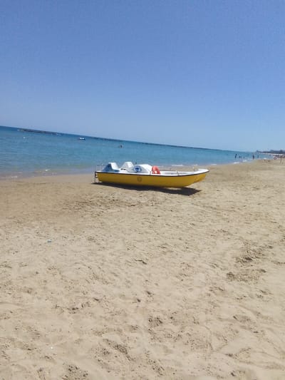 A pedal boat, yellow with white seating and a blue eye graphic, rests on a sandy beach next to the calm blue sea under a clear sky. The boat has a red life preserver attached to its side. In the distance, several people are visible in the water and on the beach, and a breakwater extends into the sea. The scene depicts a sunny day at a beach resort, likely in a coastal area of Italy.