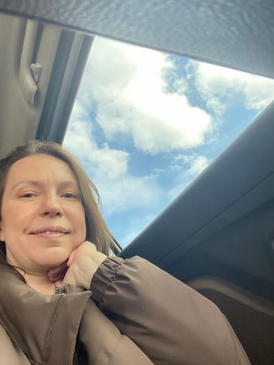 A woman is captured in a selfie-style photograph taken from inside a car, looking upwards through an open sunroof. The primary view beyond the car's interior is a bright blue sky filled with scattered white clouds, suggesting clear daytime weather.
The woman, with light brown hair, is positioned in the lower part of the frame, smiling subtly towards the camera. Her right hand is gently resting against her chin. She is wearing a brown or khaki-colored long-sleeved garment, possibly a jacket or top, which has elasticated cuffs. The car's dark interior headliner and parts of the sunroof frame are visible, along with a beige overhead grab handle on the left side of the frame. The overall impression is one of relaxation while traveling during daylight hours in Verkhnie Alnashi, Russia. No visible text is present in the image.