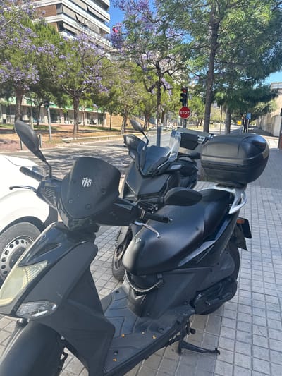 This image captures an urban street scene in La Fontsanta I Can Fatjó, Spain, on a clear, sunny day. In the immediate foreground, two black scooters are parked on a sidewalk composed of light grey square paving stones. The scooter closest to the viewer is black with a clear windscreen displaying a white, stylized "VANS" logo. It features a visible headlight, turn signals, handlebars with a chrome brake lever, and a black rearview mirror. Minor scratches are present on the front right fairing. A white car is partially visible to the left of this scooter. Behind the first scooter, a second dark-colored scooter is parked, equipped with a large black storage top case mounted on its rear rack. Its seat, a portion of its handlebars, and a black rearview mirror are visible.
In the midground, beyond the scooters and sidewalk, a paved street runs parallel to the foreground. A red traffic light with a single illuminated red signal is visible, accompanied by a circular red "STOP" sign on the same pole. Further down the street, another traffic light shows a green signal. The street is lined with numerous trees, many of which are adorned with vibrant purple blossoms, indicative of Jacaranda trees, interspersed with trees displaying green foliage. Across the street, a row of low-rise buildings houses ground-floor businesses with visible storefronts and awnings, topped by residential or commercial floors. Several indistinct human figures are visible walking on the distant sidewalk or near the street.
The background features a prominent, multi-story building with a light-colored facade and numerous windows, many equipped with dark, horizontal window blinds or awnings. The sky is clear and blue, occupying the upper portion of the frame. Additional trees and buildings are visible further in the distance under the bright daylight.