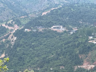 The image captures a panoramic view of a mountainous, forested landscape near the city of Uchterek, Uzbekistan. The terrain is characterized by steep, tree-covered slopes with some areas of exposed earth and rock, indicative of construction or natural erosion. Several structures are visible, including multi-story buildings and smaller houses, nestled into the hillsides. Some of these buildings appear to be under construction, with visible scaffolding and unfinished elements. A road winds through the landscape, with a white car visible on it in the lower right quadrant. Power lines and poles are also present, suggesting ongoing infrastructure development. The overall impression is one of a developing or inhabited mountainous region, possibly a resort area given the modern architecture and the presence of what might be a ski lift infrastructure on the left side of the image. The vegetation is dense and green, predominantly consisting of coniferous trees. The sky is overcast, casting a soft light over the scene.