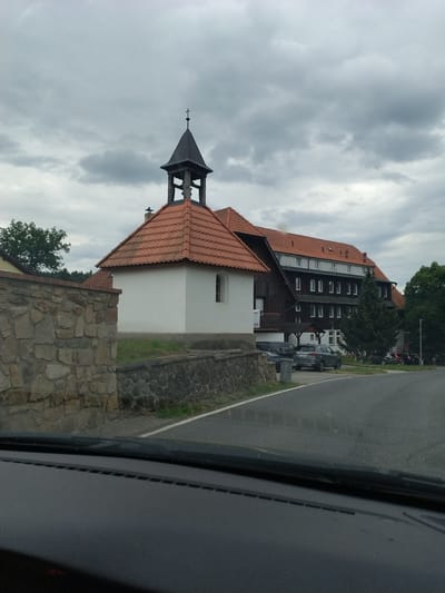 The image shows a small white chapel with a dark bell tower topped with a cross, adjacent to a larger, multi-story building with a red tiled roof. The larger building appears to be a hotel or guesthouse, with many windows visible. A stone wall runs along the left side of the image, bordering a grassy area and a road. Several cars are parked in front of the larger building. The sky is overcast with dark clouds. The scene is located in Olešná, Czechia.