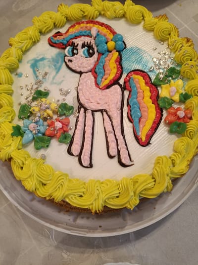 This is an image of a decorated cake with a cartoon pony figure on it, placed on a table. The cake is round and covered in white frosting, with yellow frosting piped around the edge in a decorative pattern. The pony is depicted in the center, with pink frosting for its body and a multi-colored mane and tail made of red, yellow, blue, and pink frosting. The pony has large blue eyes and small blue and yellow details on its head. There are also small, colorful frosting flowers and green leaves scattered on the cake, along with silver edible glitter. The table appears to be covered with a light-colored tablecloth. The image does not provide any information about the location in the city of Первомайское, Russia.