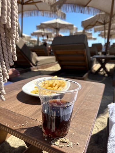The image displays a beachfront leisure scene in Ornos, Greece. In the foreground, a clear plastic cup containing a dark, carbonated beverage with visible bubbles sits on a dark brown wooden side table. Fine granular debris is scattered on the table surface around the cup's base. Adjacent to the cup, on a white plate, is a portion of potato chips. A thick, braided beige rope hangs on the left side, with a bare foot partially visible behind it. A white towel is present on the right edge of the table. The midground shows multiple wooden-framed beach lounge chairs with dark cushions, positioned on sand. Some chairs are occupied by indistinct figures. The background features numerous light-colored beach umbrellas with fringed edges, set against a clear blue sky.