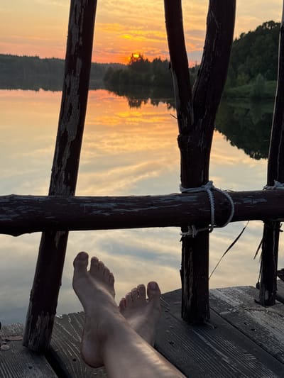 The image shows a close-up view of a person's bare feet resting on a wooden platform, with a serene sunset reflected in a body of water in the background. The wooden platform appears to be part of a dock or pier, with rustic wooden posts and a weathered railing visible. The railing is secured with rope in some areas. The sunset casts a warm glow across the sky and water, with shades of orange, yellow, and pink. The reflection shows clouds and the silhouetted outline of trees and a distant shoreline. The image is taken from a low angle, focusing on the feet and the foreground of the platform. The specific location in the city of Kamysh, Russia, is not discernible from the visual information presented in the image.