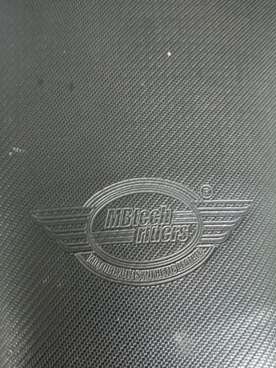 A close-up shows a textured, dark gray surface, possibly a motorcycle seat. An oval-shaped emblem with wings is embossed. The emblem features "MBtech Riders" in stylized letters within the oval. Below the oval, "MOTORCYCLE SYNTHETIC LEATHER" is inscribed in a curved, smaller font. The surface displays a woven or carbon fiber-like pattern.