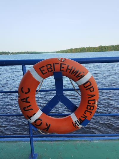 An orange life preserver with black Cyrillic lettering is mounted on a blue railing on the deck of a vessel. The life preserver reads "СПАСИ КОТЛАС ЕВГЕНИЙ ПРАВЕДНЫЙ" which translates to "SAVE KOTLAS YEVGENY RIGHTEOUS". The vessel is on a body of water with a forested shoreline in the distance. The sky is clear and light blue. The location is Bessolovo, Russia.