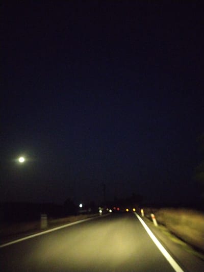 The image depicts a road at night, possibly in or near Mešno, Czechia. A bright, possibly full or near-full moon is visible in the dark sky on the left side of the frame. The road itself is illuminated by headlights, casting a yellow glow on the asphalt. White lines mark the edges of the road, and a reflective post with red and white markings is visible on the right side. In the distance, other vehicle lights and possibly streetlights create blurry orbs of light. The surrounding environment appears to be rural, with dark, indistinct shapes suggesting fields or trees along the roadside. The overall impression is one of driving on a quiet road under a night sky.