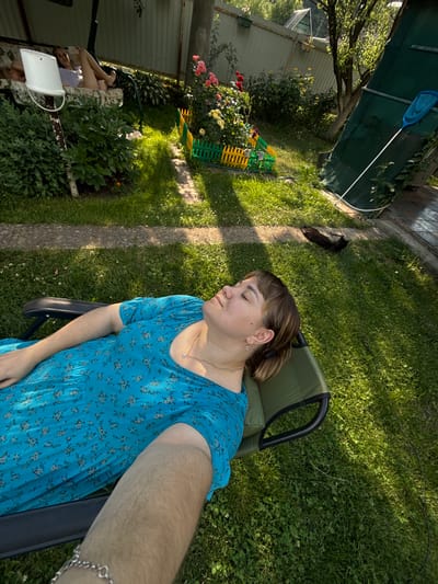 In an outdoor setting, a woman with light brown hair tied in a bun is resting in a green lounge chair. She is wearing a light blue dress with a floral pattern. Her eyes are closed, and she appears to be relaxed and sleeping, with a gentle smile on her face. A silver bracelet is visible on her wrist. The scene is bathed in the warm sunlight of what appears to be a late afternoon or early evening.
The background shows a lush green lawn with scattered white clover flowers. A stone pathway runs diagonally across the grass. To the left, there are rose bushes with pink and yellow flowers, and a colorful plastic fence. Behind the flowers, a light-colored fence and a dark green tarpaulin are visible, suggesting a backyard or garden area. In the distance, trees and foliage can be seen. A black cat is lying on the grass near the woman, also seemingly resting.
The overall impression is one of peaceful relaxation in a garden on a pleasant day. The location context provided is Nizhne-Chufichevo, Russia. There is no visible text in the image.