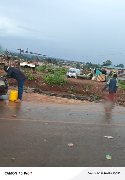 This image captures a street scene in Gitaru ward, Kenya, under an overcast sky. The foreground features a wet asphalt road, indicating rain. On the left, a male subject in a black long-sleeved shirt, blue jeans, and light brown shoes is bent over the front of a light brown vehicle, appearing to interact with its engine compartment. A yellow jerrycan rests on the ground next to his left foot. On the right, a second male subject, wearing a blue and grey jacket with red pants, walks away from the viewer on the shoulder of the wet road, kicking up water or mud.
The middle ground is an elevated, reddish-brown dirt embankment leading up from the road. This area hosts multiple rudimentary market stalls or shelters constructed from wooden poles and various coverings such as tarpaulins or canvas. Green shrubbery is interspersed on the slope. Further up the embankment, a silver van (KAE 577J) is parked, partially covered by a white sheet. Adjacent to the van is a makeshift structure built from corrugated metal and other materials, with wooden pallets stacked nearby.
The background reveals a gently sloping landscape with numerous scattered residential and commercial buildings, characteristic of a semi-urban settlement. A two-story building with a distinctive blue roof is visible in the far right background. Overhead, utility lines traverse the scene. The overall environment suggests a bustling, developing area.