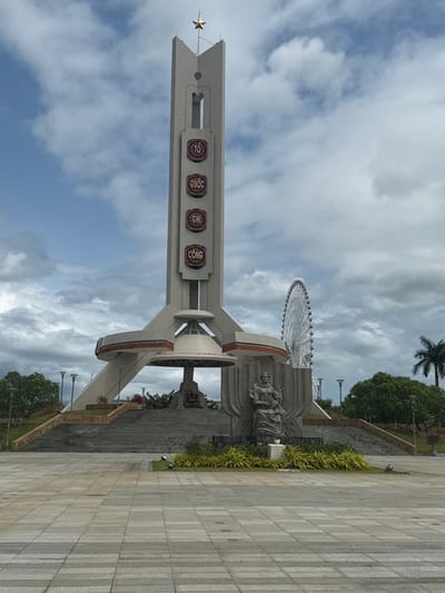 The image depicts a monumental plaza in Hòa Cường Ward, Vietnam, under an overcast sky. The central feature is a tall, light-grey monument with a rectangular column topped by a golden five-pointed star. Vertically aligned on the column are four circular plaques bearing the Vietnamese text "TỔ QUỐC GHI CÔNG" (The Fatherland Recognizes the Merit). The monument's base is broad and angular, featuring a central inverted saucer-shaped structure. Dark stone steps lead up to the base of the monument. To the right of the steps (viewer's left), a dark grey statue of a seated woman holding a child is positioned in front of a stylized backdrop. The foreground consists of a wide, light-grey paved plaza with a grid pattern. In the mid-ground, a large white Ferris wheel is visible to the right of the main monument. Scattered green landscaping, including low bushes in planters and mature trees, borders the plaza and monument. Several modern streetlights are visible throughout the scene. No individuals are present in the image.