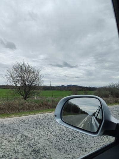 The image depicts a view from inside a moving vehicle, likely a car, on a road near Ohoden, Bulgaria. The road is asphalt with some visible wear. In the foreground, the passenger side mirror reflects the road behind the vehicle. Through the front windshield, a landscape is visible with a green field, a bare tree, shrubs, power lines, and a distant mountain under an overcast sky. There are no visible people.