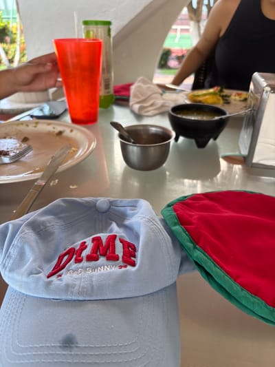 An overhead shot captures a cluttered tabletop. A light blue baseball cap with red embroidered lettering sits next to a red and green potholder. Nearby, there's a dirty plate with utensils, an orange plastic cup, and a metal bowl with a spoon. A black pot and a container with a green lid are also present. The background reveals a partial view of a person's arm and a glimpse of an outdoor scene.