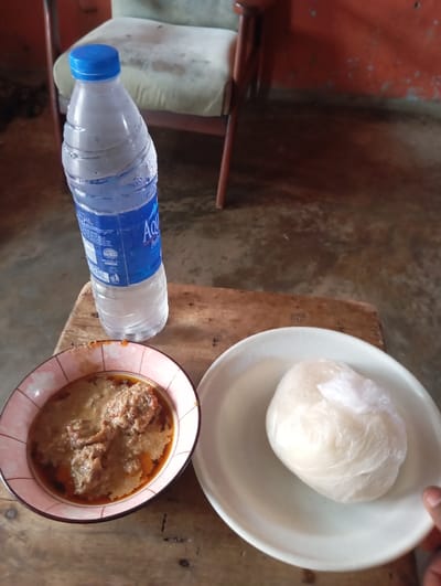 The scene, set in Okere-Urhobo, Nigeria, features a meal resting on a light-brown wooden surface. A clear plastic bottle of Aqua Rapha Purified Water with a blue cap shows condensation. Adjacent to it, a pink-patterned ceramic bowl contains a light-brown stew with protein chunks and a reddish oil layer. A white circular plate holds a white, rounded mass of "swallow" food, encased in clear plastic film. A dark-skinned human hand is partially visible at the bottom right, holding the edge of the white plate. In the background, a worn armchair with light-colored upholstery and wooden arms stands against a reddish-orange wall exhibiting wear and discoloration. The floor appears to be a discolored, uneven surface.