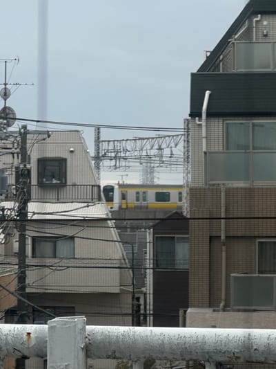 Buildings with windows and varying heights fill the frame, along with utility poles and wires. A glimpse of a train, mostly yellow with some white, is visible between the buildings. A white, cylindrical structure occupies the right foreground. The sky is overcast and pale, adding to the muted color palette.