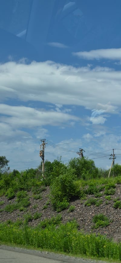 The image shows a roadside view with a blue sky filled with white clouds. In the middle ground, there is a steep, rocky slope covered with green vegetation, including trees and bushes. Several utility poles with electrical wires and transformers are visible along the top of the slope. The foreground shows a paved road surface with some grass and low-lying plants at the edge. The reflection of what appears to be a vehicle's interior or structure is visible on the upper part of the image, suggesting the photo was taken from inside a moving vehicle. The specific location in the city of Gorodechnoe, Russia, is not identifiable from the visual elements in the image alone.