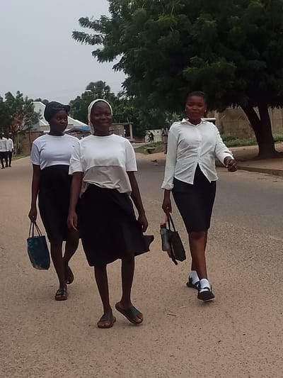 Three young African women are walking on a paved road in Biliri, Nigeria. They wear similar uniforms of white shirts and black skirts, each carrying a bag. The woman on the right wears white socks with her sandals and an unbuttoned long-sleeve shirt. The other two women wear open sandals. A tree provides shade in the background and buildings are visible.