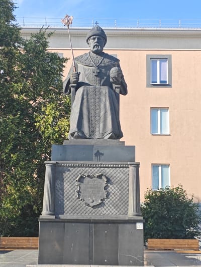 A monumental statue of a seated male figure in religious vestments stands prominently in an urban setting. The figure, rendered in dark metal, is adorned with a jeweled crown, a cross on its chest, and holds a scepter in its right hand and an orb in its left. The statue is erected on a multi-tiered, dark stone pedestal. The pedestal features decorative relief work, including a coat of arms or crest at its front. Behind the statue, a light-colored building with several windows is visible, suggesting a public space or administrative area. Trees with green foliage flank the statue and the building. The sky above is clear and blue. A wooden bench is positioned in the foreground. This is a statue of Metropolitan Peter of Moscow, located in Novyy Oskol, Russia.