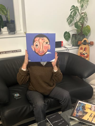 A person reclines on a black couch, holding a vinyl record depicting a cartoonish face, obscuring their own. They wear a brown sweater and gray pants. A skateboard and a potted monstera plant are on the wall behind the couch. A Nintendo Switch rests next to the plant. Two books, including one titled "No Rain, No Flowers," and a phone lie nearby on the couch. A decorative sign reading "SLEEP NUMBER IS 4" is visible near the window.