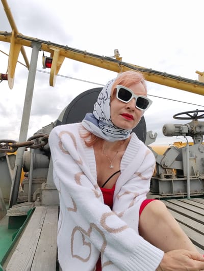 A woman with pink hair, wearing a white cardigan with heart patterns, a red top, a headscarf, and white-framed sunglasses, poses for a photo on a boat. She is seated on a wooden deck, with her legs crossed. In the background, there are industrial elements of the boat, including chains, machinery, and a yellow structure. The sky is overcast. The location is identified as Novokemskii, Russia, specifically on a riverboat or ferry.
