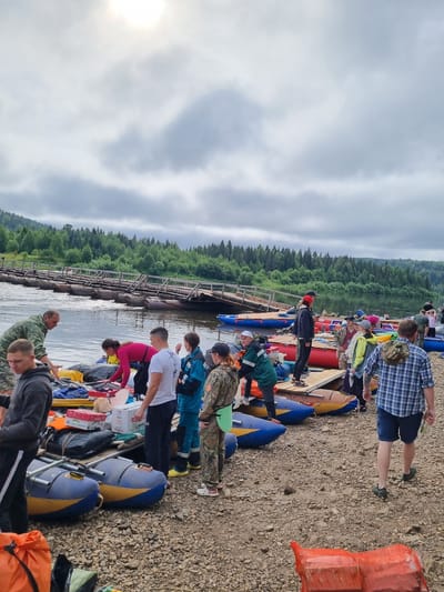 A group of people are gathered on a rocky shore next to a river, preparing for a rafting trip. In the background, a long wooden bridge spans the river, supported by inflatable pontoons. The sky is overcast, with the sun partially visible through the clouds.
There are many inflatable rafts on the shore, some of which are blue and yellow, others red and yellow. Various bags, boxes, and supplies are being loaded onto or unloaded from these rafts. Several individuals are actively engaged in this process, while others appear to be waiting or observing. The people are dressed in a mix of casual clothing and what appear to be specialized outdoor gear, including life jackets.
The environment is natural, with a dense forest visible on the far bank of the river and extending up the hillsides. The river itself appears calm. The overall impression is of a group preparing for an organized outdoor adventure activity. The image depicts a scene consistent with an outdoor recreation area near a body of water, possibly a designated launch point for river activities.