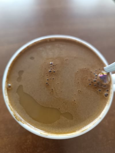 A top-down, close-up shot shows a white mug filled with coffee. The coffee has a light brown foam with several small bubbles on the surface. A portion of a white spoon is visible on the right side, partially submerged in the coffee. The mug rests on a brown, textured surface, possibly a table. The lighting is soft, creating a gentle reflection on the coffee and the mug's rim. The location is identified as Skidzieĺ, Belarus, but no specific landmarks or contextual elements of that location are visible in the image.
