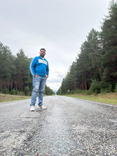 A man stands on a paved road. He is wearing jeans, white sneakers with orange accents, and a blue jacket with white stripes. The road is bordered by trees. The location is Сизая, Russia. The sky is overcast.