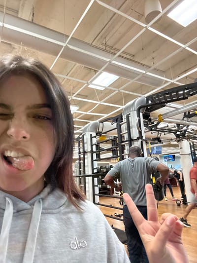 The image depicts the interior of a fitness center, specifically identified as Life Time Athletic - Plainsboro in Plainsboro Township, New Jersey, United States.

In the immediate foreground, the upper torso and head of a young female are visible, positioned for a selfie. She has dark hair, wears a grey hooded sweatshirt with an "alo" logo, squints her eyes, and extends her tongue from her mouth. Her left hand is raised, displaying a two-finger peace sign.

The background reveals the gym floor, populated by multiple individuals and exercise equipment. A muscular male, seen from behind, wears a grey t-shirt and dark pants, holding black straps attached to a large grey and black multi-station fitness rig with yellow accents. A white sign on the equipment includes the text "OUTRACK." Other gym patrons are present, including one near a red kettlebell and another partially visible in an orange shirt. A blue display screen is discernible in the distance. The flooring is a light wood finish. The ceiling is a grid-style drop ceiling with recessed rectangular fluorescent lights, suspended circular pendant lights, and visible grey and white cylindrical air ducts.