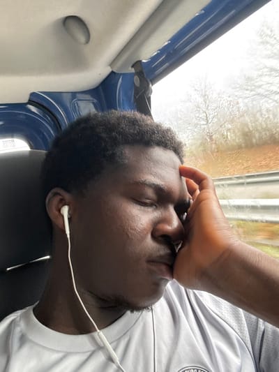 A dark-skinned male individual is depicted seated within a moving vehicle in Dettwiller, France. He has short, dark, curly hair, and his eyes are closed. His left hand is raised, with fingers resting on his forehead and his palm supporting his left cheek. He is wearing white wired earphones in both ears, with the wires visible against his light grey, short-sleeved shirt, which features dark grey patterned shoulders and a partial logo on the chest. The vehicle's interior shows a dark blue paneling above the window and a light-colored ceiling with an oval light fixture. Through the window on the right, a blurred exterior environment is visible, featuring bare trees against an overcast sky, brown and orange ground foliage, and a metal guardrail, indicative of travel on a road or highway.