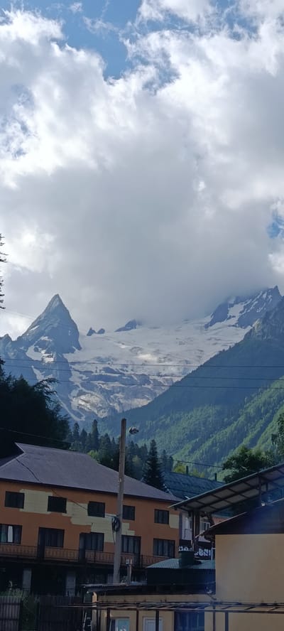 The image shows a dramatic mountain landscape in Dombay, Russia, under a partly cloudy sky. Dominating the background are snow-capped peaks and glaciers, with a sharply pointed mountain cone to the left, partially obscured by clouds. The midground features a dense, green forest covering the mountain slopes. In the foreground, several buildings are visible. On the left, a multi-story building with an orange and cream facade and dark-paned windows is prominent. To its right, a ski lift with a red gondola is suspended in the air, passing by a utility pole. More buildings with dark roofs and some covered outdoor structures are present in the lower right portion of the frame. The overall scene captures a blend of natural beauty and human infrastructure in a mountainous resort area.