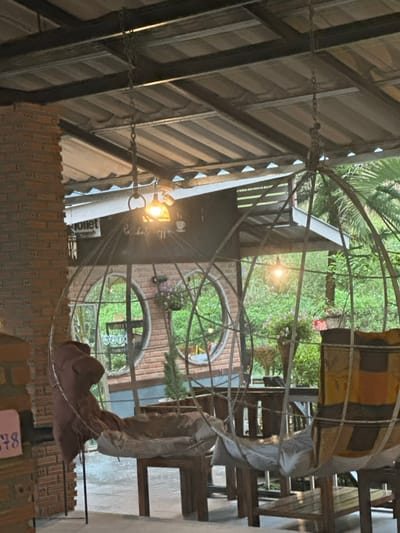 The image depicts an unoccupied semi-outdoor seating area in Ban Paem Bok, Thailand. The scene is illuminated by artificial lights suspended from a corrugated metal roof supported by dark metal beams. Two spherical, open-cage metal hanging chairs dominate the foreground, each suspended by chains. The chair on the left contains a brown cushion and light fabric, while the chair on the right features a multi-colored striped cushion (yellow, brown, black) and light fabric. Wooden tables and chairs are arranged between and beyond these hanging chairs. In the mid-ground, a brick building with two large circular window-like openings is visible; these openings contain hanging baskets of purple flowers. Above the building's entrance, a white sign displays the word "Toilet" and "Coffee" alongside a coffee cup icon. A partial brick column is visible on the far left, and lush green foliage and trees form the background. No individuals are present within the frame.
