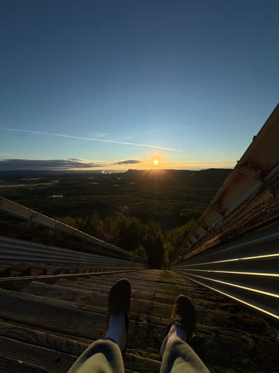 The image depicts a first-person perspective of a person sitting on the wooden ramp of a ski jump in Oliver Paipoonge, Canada, at sunrise. The sun is low on the horizon, casting long shadows and illuminating the landscape. The ski jump ramp is constructed of weathered wooden planks and metal sheeting, with some patches of moss growing on the wooden sections. The ramp descends steeply into a valley covered in dense green forest. In the distance, the land flattens out, with a body of water visible and faint plumes of smoke rising from what appear to be industrial buildings. The sky is a clear blue, with a thin contrail from an airplane streaking across it. The person's legs are visible in the foreground, wearing green pants and dark shoes, with white socks. The overall scene conveys a sense of tranquility and expansive natural beauty at the start of a new day.