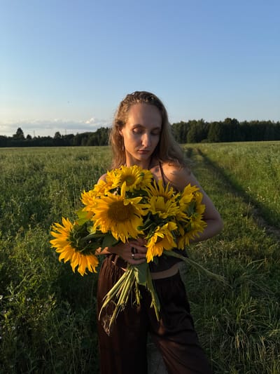 In a field with tall green grass, a young woman with blonde hair holds a large bouquet of sunflowers. She is wearing a dark brown crop top and pants. The woman's eyes are closed, and her head is slightly bowed. The background shows a dirt path leading into a forest of dark green trees under a clear blue sky with a few white clouds. The lighting suggests it is late afternoon or early evening. This scene is located in the city of Aksaitovo, Russia.