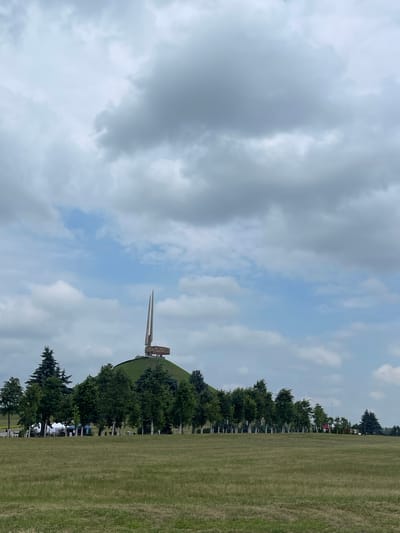 This image captures an outdoor scene with a prominent monument in the background. The monument appears to be a tall, slender structure, possibly a sword or obelisk, rising from a green, sloped hill. Trees are planted in a line along the base of the hill, leading towards the monument. In the foreground, a large expanse of mowed grass stretches to the right side of the frame, with more trees and some indistinct objects or people visible further in the distance along a road or pathway. The sky above is filled with a mix of gray and white clouds, suggesting a partially overcast or changeable weather day. The orientation of the image is vertical, with the sky occupying the left portion and the landscape filling the right. The overall atmosphere appears to be calm and potentially commemorative, given the nature of the monument. There is no visible text in the image.