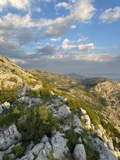 This image depicts a mountainous landscape in Općina Tučepi, Croatia, under a sky filled with scattered clouds. In the foreground, a winding asphalt road is visible, bordered by rocky terrain and sparse green vegetation. The road ascends a hillside, revealing a dramatic drop-off to the right. Beyond the immediate foreground, the landscape unfolds into a series of rolling hills and rugged mountains, dotted with patches of greenery. In the distance, a body of water, likely the Adriatic Sea, is faintly visible, with islands or peninsulas silhouetted against the horizon. The sunlight, casting long shadows, suggests it is either early morning or late afternoon. There are no people or animals present in the scene. The environment is characterized by its natural, untamed beauty, with rocky outcrops, scrubby bushes, and hardy grasses dominating the terrain.