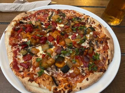 A full, circular pizza, approximately 12 inches in diameter, is centered on a white ceramic plate, which rests on a wooden table composed of horizontal planks. The pizza has a golden-brown crust with visible charring and is topped with red tomato sauce, melted white cheese, and a generous assortment of diced vegetables including red bell peppers, orange/yellow bell peppers, green zucchini, and sliced mushrooms. Small green microgreens are scattered over the toppings. To the upper left of the plate, a crumpled light brown paper napkin is visible on the wooden surface. To the upper right, a partial view of a clear glass containing an amber-colored liquid, consistent with beer, is present. No individuals are depicted, and no specific actions or interactions are occurring. The precise location within Dorchester, United Kingdom, cannot be determined from the visual information provided.