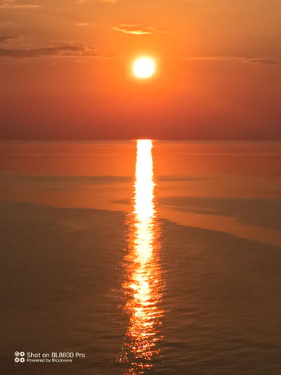 The image captures a serene sunset over the Black Sea, with the sun positioned above the horizon, casting a vibrant orange glow across the sky and water. A bright reflection of the sun streaks across the calm sea surface, creating a shimmering path of light. The lower portion of the image displays a watermark indicating "Shot on BL8800 Pro" and "Powered by Blackview". The scene appears to be located near Kamen Bryag, Bulgaria, known for its stunning coastal sunsets. There are no people or man-made structures visible in the frame, emphasizing the natural beauty of the environment.