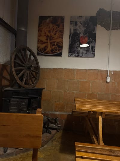 The image depicts an interior scene, possibly a restaurant or a decorative space in Chipilo de Francisco Javier Mina (Chipilo), Mexico. On the left wall are two framed pictures: one featuring a still life with what appears to be wine glasses and a red object, and another showcasing pasta. Below the pasta picture, a gray pipe is visible against the wall.
The right side of the image shows a brown brick wall. Adjacent to the brick wall is a wooden slat structure, potentially part of a seating arrangement. A dark-colored cabinet stands next to a wooden wheel. On top of the cabinet, there is an old-fashioned appliance, possibly a grinder, and a portion of a wooden table. The floor appears to be uneven and made of a textured material.