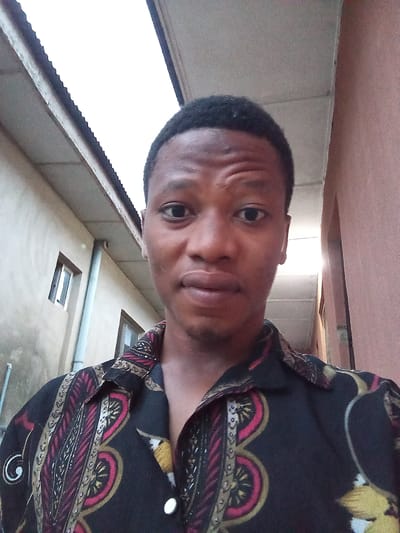 A young Black male, appearing to be in his late teens or early twenties, poses for a selfie in an outdoor urban setting in Oshogbo, Nigeria. He has short black hair and is looking directly at the camera with a neutral expression. He is wearing a dark-colored short-sleeved shirt with a vibrant, intricate pattern featuring shades of red, yellow, and gold. The background reveals architectural elements, including a textured, light-colored wall with a window, a downspout, and an overhanging corrugated metal roof. To the right, a reddish-brown wall is visible, suggesting the presence of adjacent buildings. The lighting indicates it is daytime, possibly late afternoon, with some shadows visible.