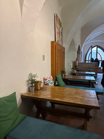 The image depicts the interior of a dining establishment in the city of Znojmo, Czechia. The room features white, textured walls with visible signs of wear and a vaulted ceiling supported by arched structures. The floor is tiled with reddish-brown square pavers.

In the foreground and extending into the midground, multiple rustic wooden tables with trestle bases are arranged. Each table is accompanied by long wooden benches fitted with teal blue cushions. Green throw pillows are placed on the benches and against the wall. On the nearest table, a small potted plant in a silver bucket, a wooden block holder, a salt and pepper shaker, and a coaster with "BOHÉM" printed on it are visible. A multi-colored cushion with a geometric pattern rests on a bench slightly further back.

Against the right wall, a tall wooden cabinet with two doors, each carved with a chevron or zigzag pattern, stands. Above this cabinet, a large, colorful abstract artwork is mounted on the wall. Further into the background, additional tables and benches are arranged. At the far end of the room, a large arched opening, likely an entrance or exit, allows natural light to enter. Several individuals are partially visible near this opening, appearing as silhouettes or dark figures, some wearing coats, suggesting they are either entering, exiting, or standing near the threshold. Their specific actions or interactions are not discernible.