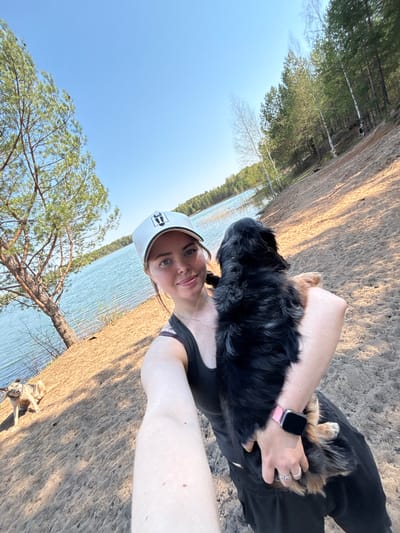The image depicts a woman holding a small dog, likely a Bernese Mountain Dog puppy, outdoors near a body of water. The woman wears a white cap, a dark tank top, and a pink smartwatch. She is facing the camera and smiling. The dog is mostly black with some brown markings, held close to her chest. The background includes a shoreline with sandy ground, trees, and the surface of a lake or wide river. The sky is blue. The location appears to be a natural setting, but there is no specific evidence to confirm it is in Selychka, Russia. There is no evidence of urban development in the immediate vicinity. Another dog is visible in the bottom right corner of the image.