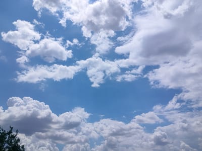 The image shows a sky with scattered white and gray clouds against a clear blue sky. The clouds vary in size and density, with some appearing fluffy and white, while others are larger and show darker gray undersides, suggesting potential depth or shadow. A small portion of green foliage is visible in the bottom left corner, indicating the presence of trees or other vegetation. The scene is static and depicts a natural outdoor environment. There is no evidence to suggest this image was taken in the city of Salsk, Russia.